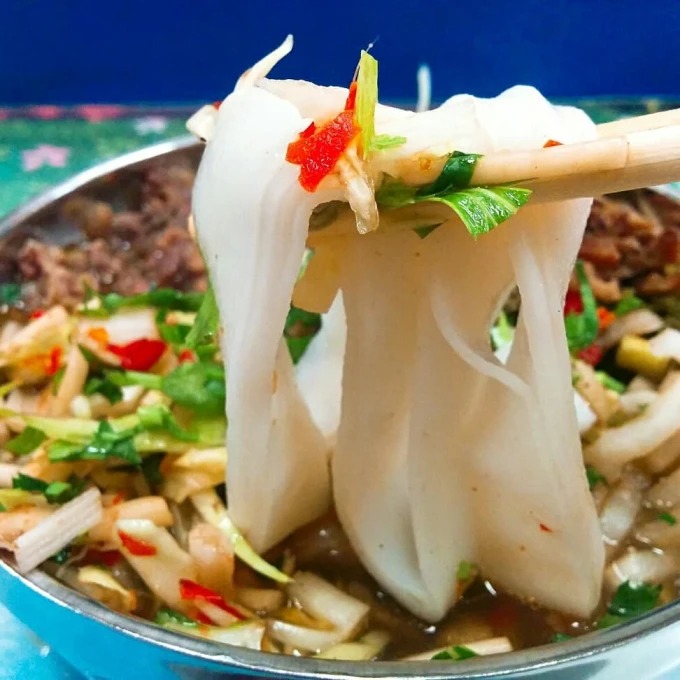 Larges nouilles de riz soulevées au-dessus d’un bol d’herbes et de piment